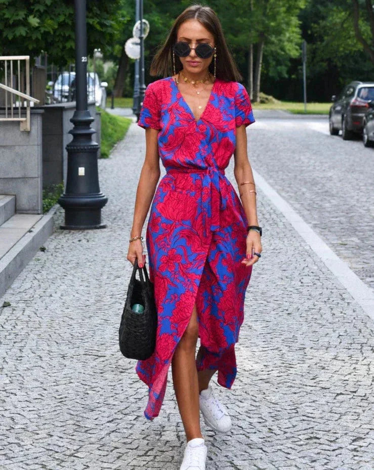 Donna con abito floreale rosso e blu, occhiali da sole, borsa nera, cammina su strada acciottolata. Moda estiva, look casual chic, tendenze 2023.