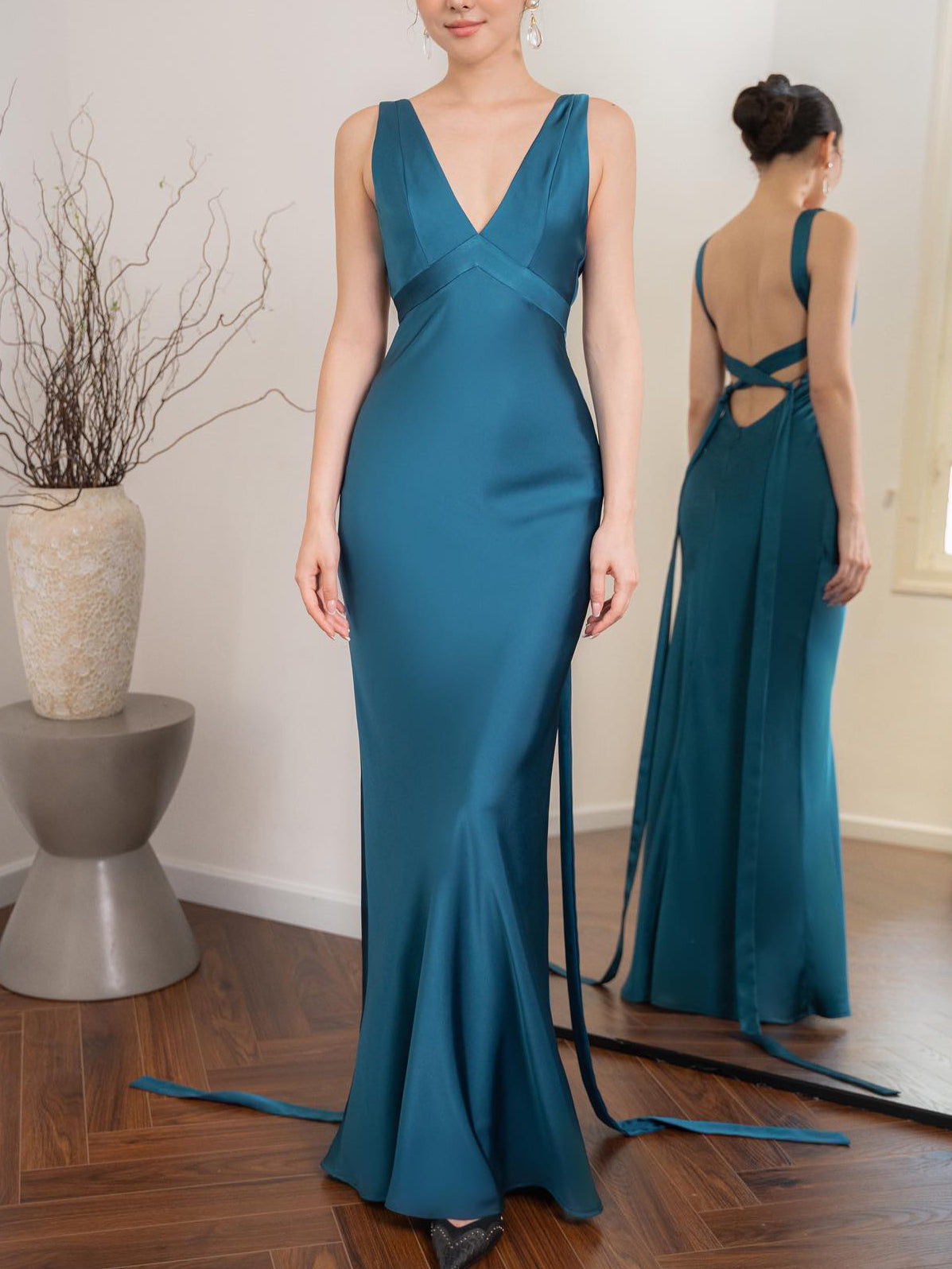 Abito da sera lungo blu in raso con scollo a V, dettagli incrociati sulla schiena, indossato da una donna in una stanza moderna con specchio.