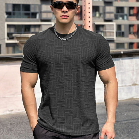 Giovane uomo muscoloso con occhiali da sole, indossa maglietta nera a trama geometrica e catena argentata, sfondo urbano moderno.