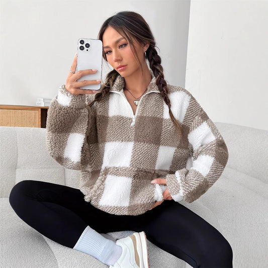 Donna con maglione a quadri beige e bianchi, seduta su divano, scatta selfie con smartphone. Look casual, moda autunnale, capelli intrecciati.