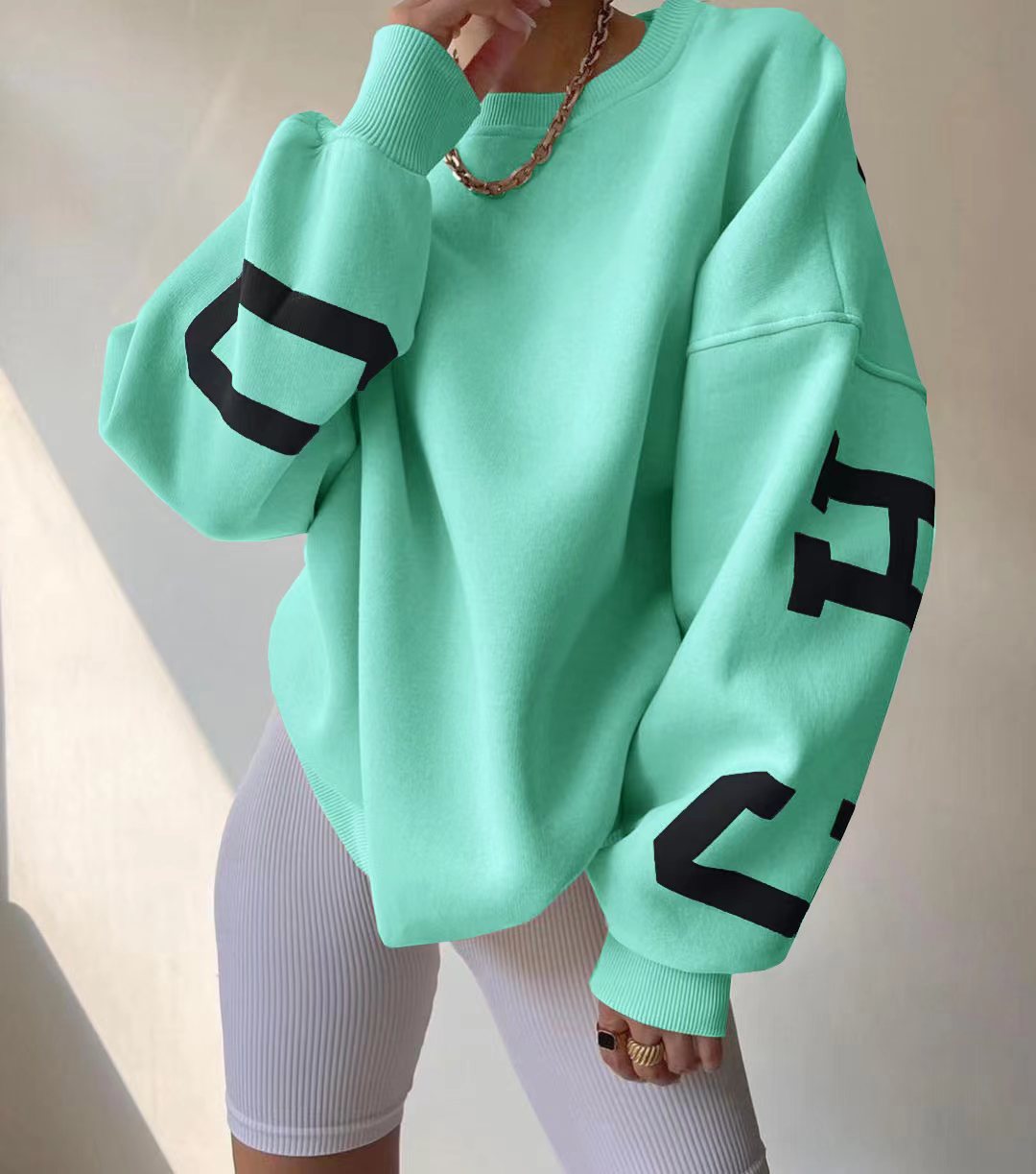 Felpa oversize verde menta con scritte nere sulle maniche, indossata con pantaloncini bianchi a coste. Stile casual e moderno per moda urbana.