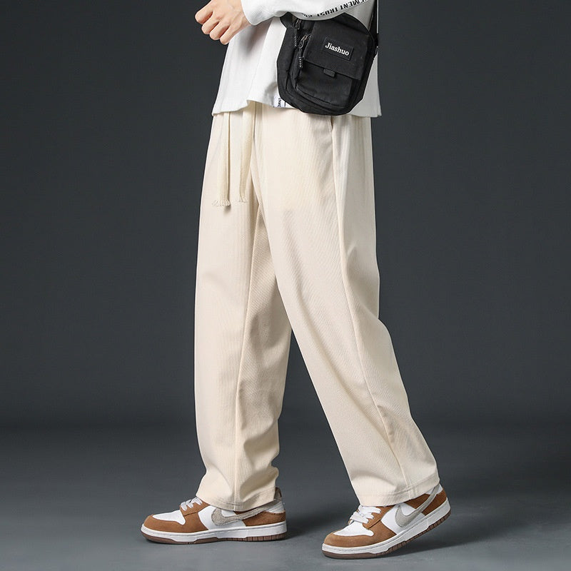 Pantaloni larghi beige uomo, abbinati a sneakers bianche e marroni, borsa a tracolla nera. Moda casual, streetwear maschile.