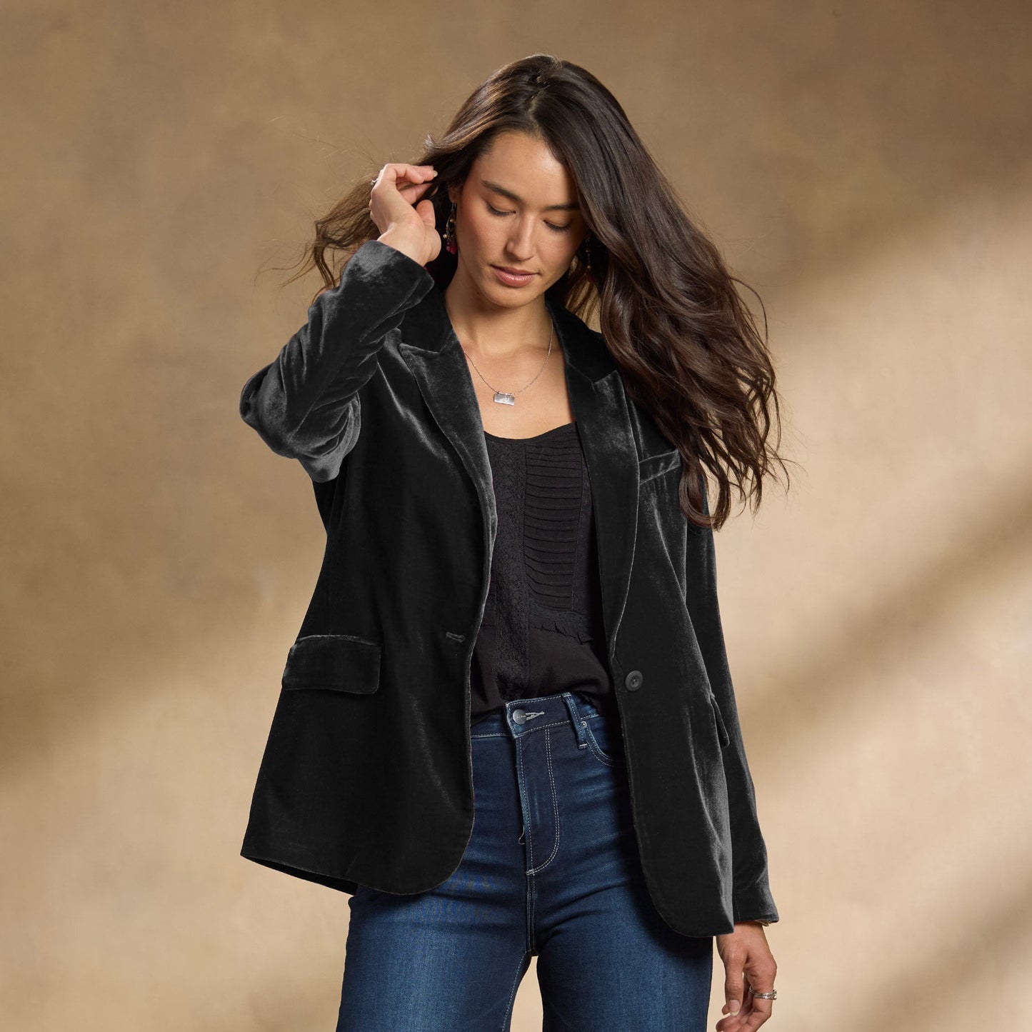 Donna con giacca di velluto nera, maglia nera e jeans blu. Moda autunno, abbigliamento elegante casual, sfondo beige.