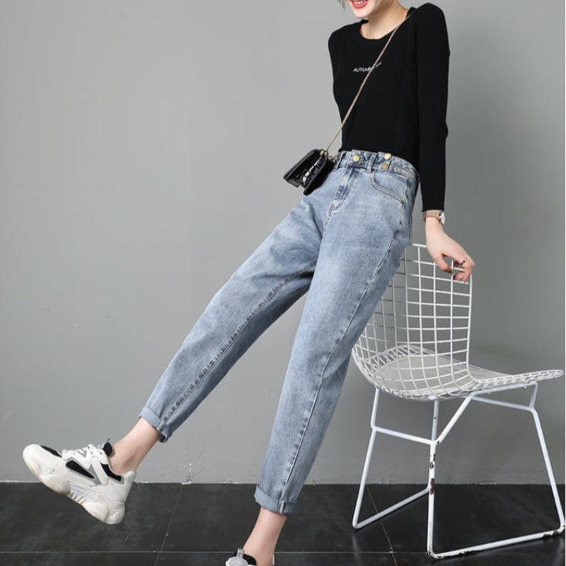 Donna in jeans boyfriend e maglia nera, seduta su sedia bianca, con borsa a tracolla e sneakers bianche. Moda casual, stile urbano.