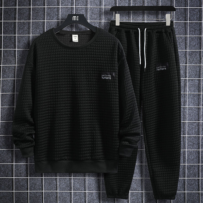 Completo tuta uomo nero, tessuto trapuntato, maglia a maniche lunghe e pantaloni con coulisse, appesi su gruccia, sfondo a griglia.