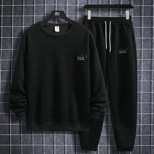 Completo tuta uomo nero, tessuto trapuntato, maglia a maniche lunghe e pantaloni con coulisse, appesi su gruccia, sfondo a griglia.
