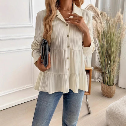 Donna in camicia bianca plissettata e jeans, tiene una borsa nera. Sfondo con piante decorative e parete bianca. Moda casual elegante.