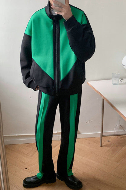 Uomo in tuta sportiva verde e nera, con giacca a zip e pantaloni abbinati, in piedi su pavimento in legno. Moda streetwear moderna.