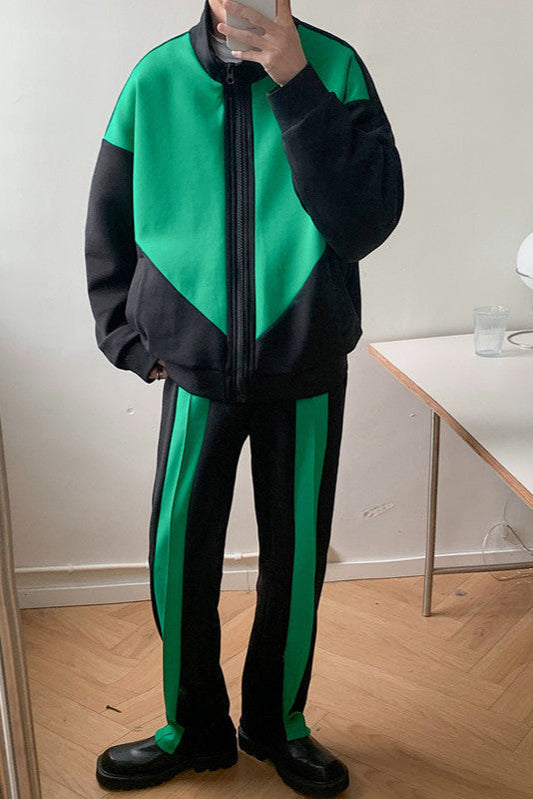 Uomo in tuta sportiva verde e nera, con giacca a zip e pantaloni abbinati, in piedi su pavimento in legno. Moda streetwear moderna.