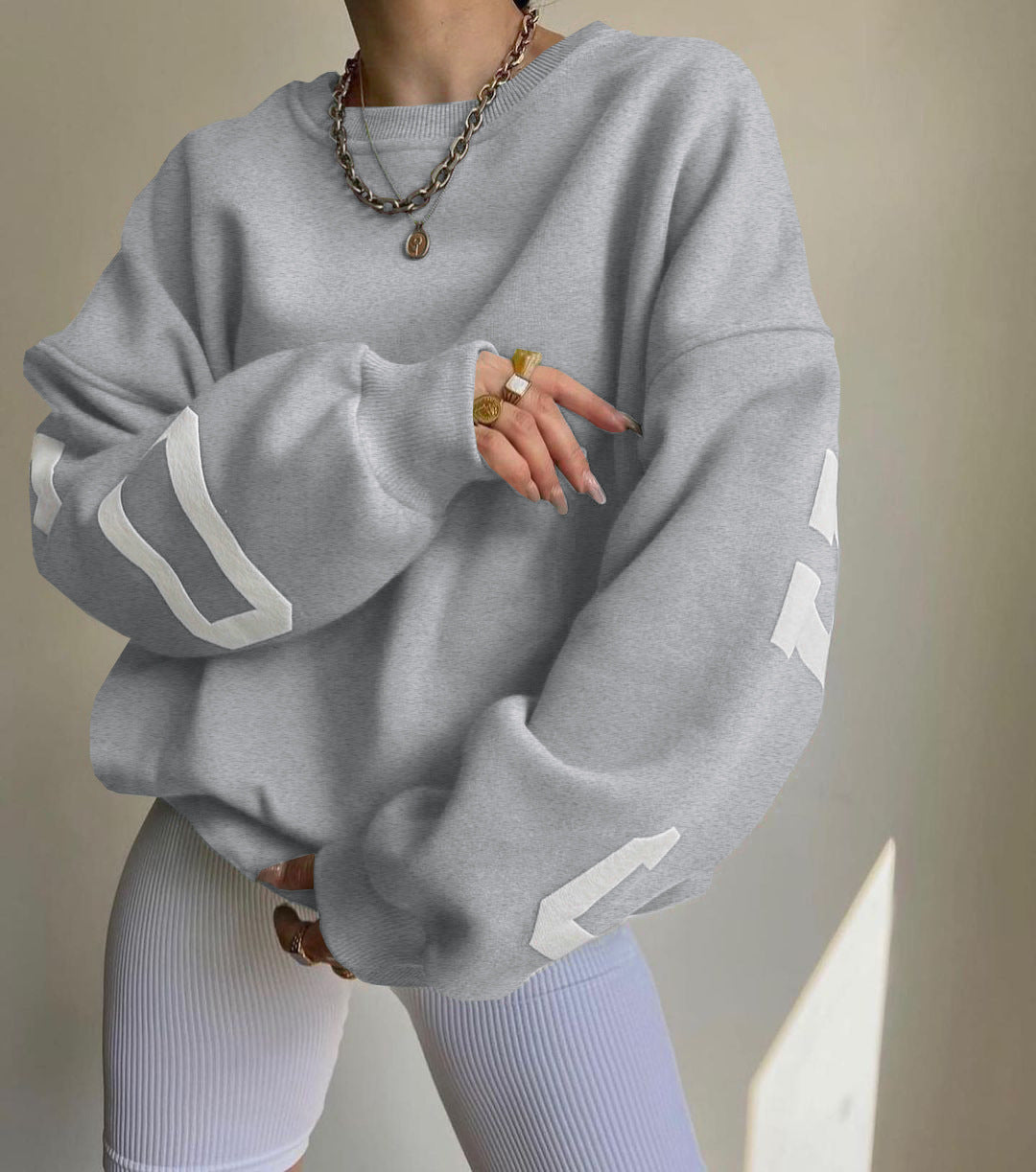 Donna con felpa grigia oversize e pantaloni a coste bianchi, indossa collana e anelli dorati. Moda casual, abbigliamento streetwear trendy.