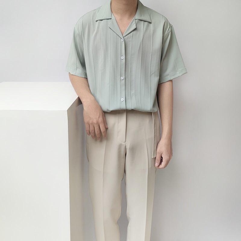 Uomo in camicia verde chiaro a righe verticali e pantaloni beige, posa accanto a un cubo bianco. Moda estiva, abbigliamento casual elegante.