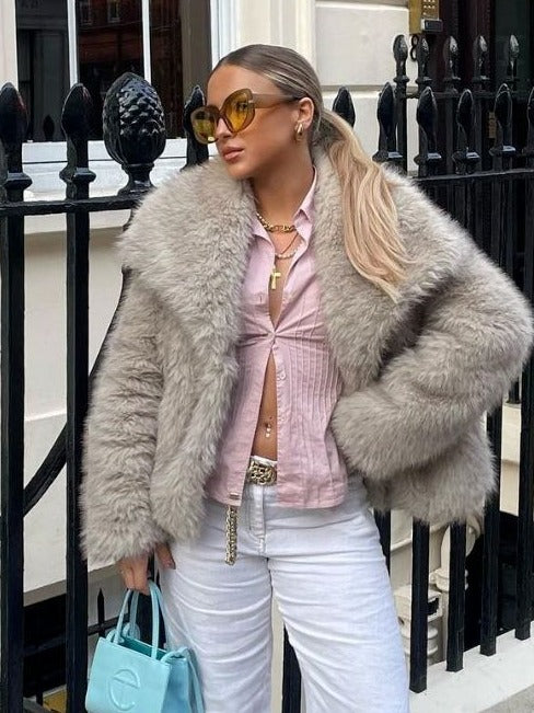 Donna con cappotto di pelliccia grigia, occhiali da sole gialli, camicia rosa e pantaloni bianchi, tiene una borsa azzurra. Moda urbana elegante.