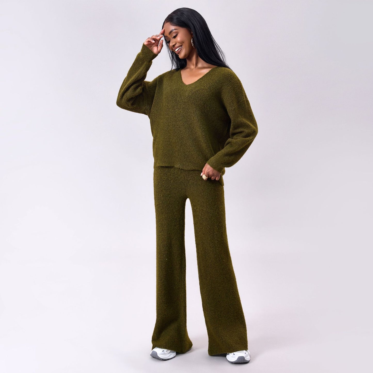 Donna sorridente in completo maglia verde oliva, maglione e pantaloni larghi, posa su sfondo grigio. Moda autunnale, abbigliamento casual elegante.