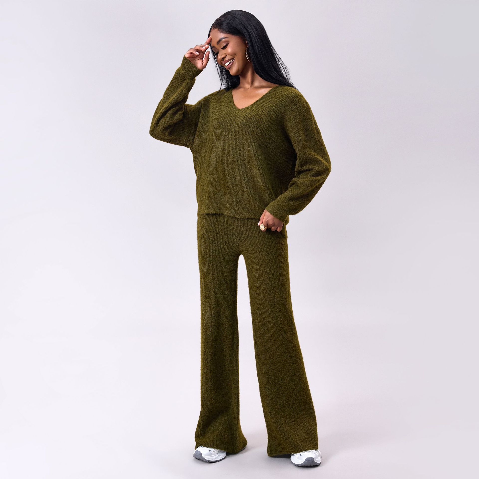 Donna sorridente in completo maglia verde oliva, maglione e pantaloni larghi, posa su sfondo grigio. Moda autunnale, abbigliamento casual elegante.