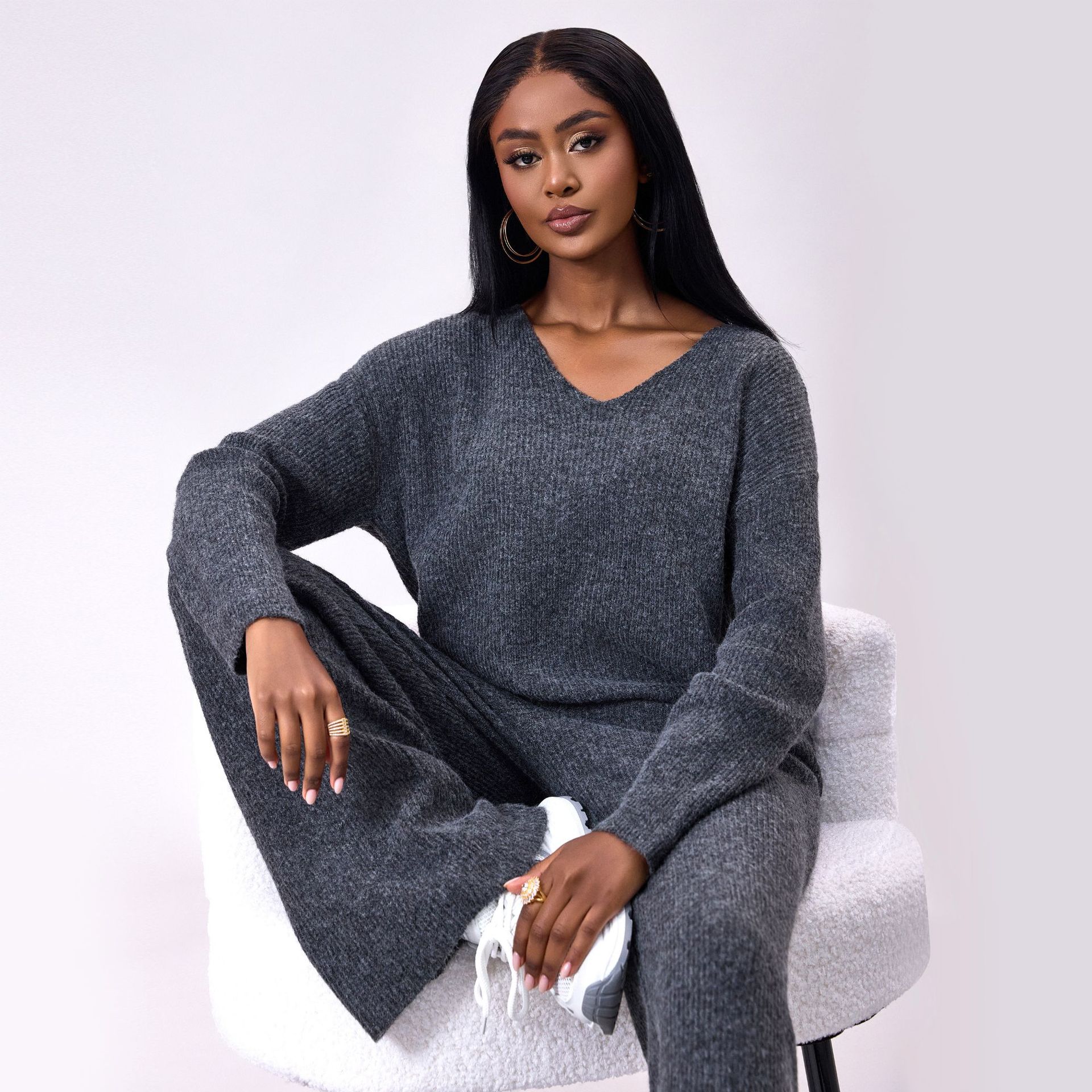 Donna seduta su sedia bianca, indossa maglione e pantaloni grigi in maglia, scarpe bianche, capelli lunghi neri, orecchini a cerchio. Moda autunnale.