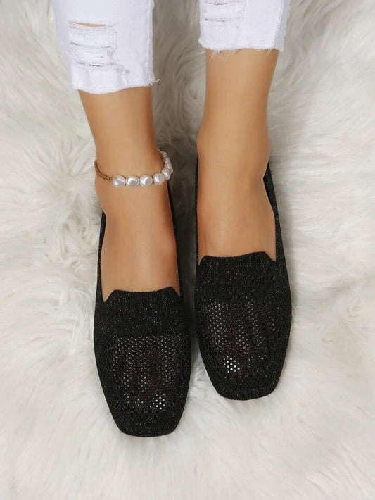 Scarpe ballerine nere scintillanti su tappeto bianco, indossate con jeans bianchi strappati e cavigliera di perle. Moda donna elegante e casual.