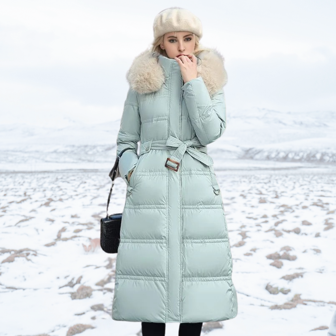 Donna in cappotto invernale verde menta con cappuccio in pelliccia, berretto beige, borsa nera, in paesaggio innevato. Moda invernale elegante.