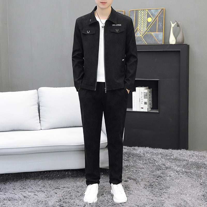 Uomo in tuta sportiva nera, giacca con zip e pantaloni abbinati, in piedi su tappeto grigio, sfondo con divano bianco e decorazioni moderne.