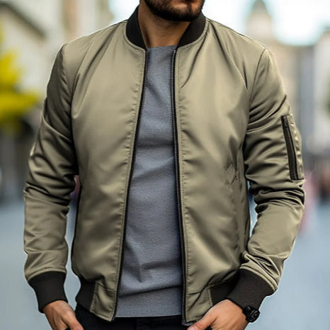 Giacca bomber verde oliva su uomo, abbinata a maglia grigia. Moda urbana maschile, stile casual autunnale. Sfondo sfocato di città.