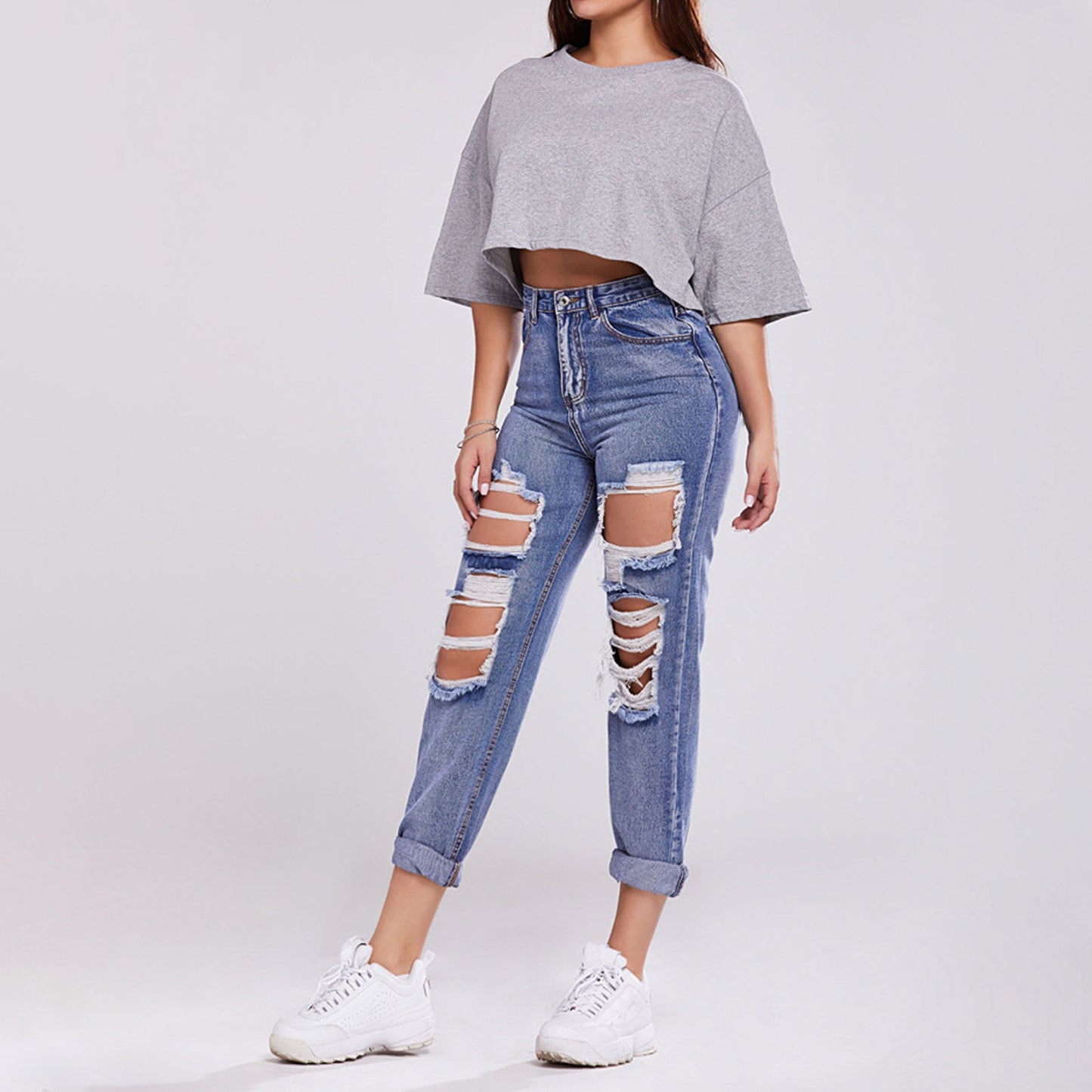 Donna in jeans strappati e maglietta grigia corta, scarpe da ginnastica bianche. Moda casual, abbigliamento trendy, stile urbano.