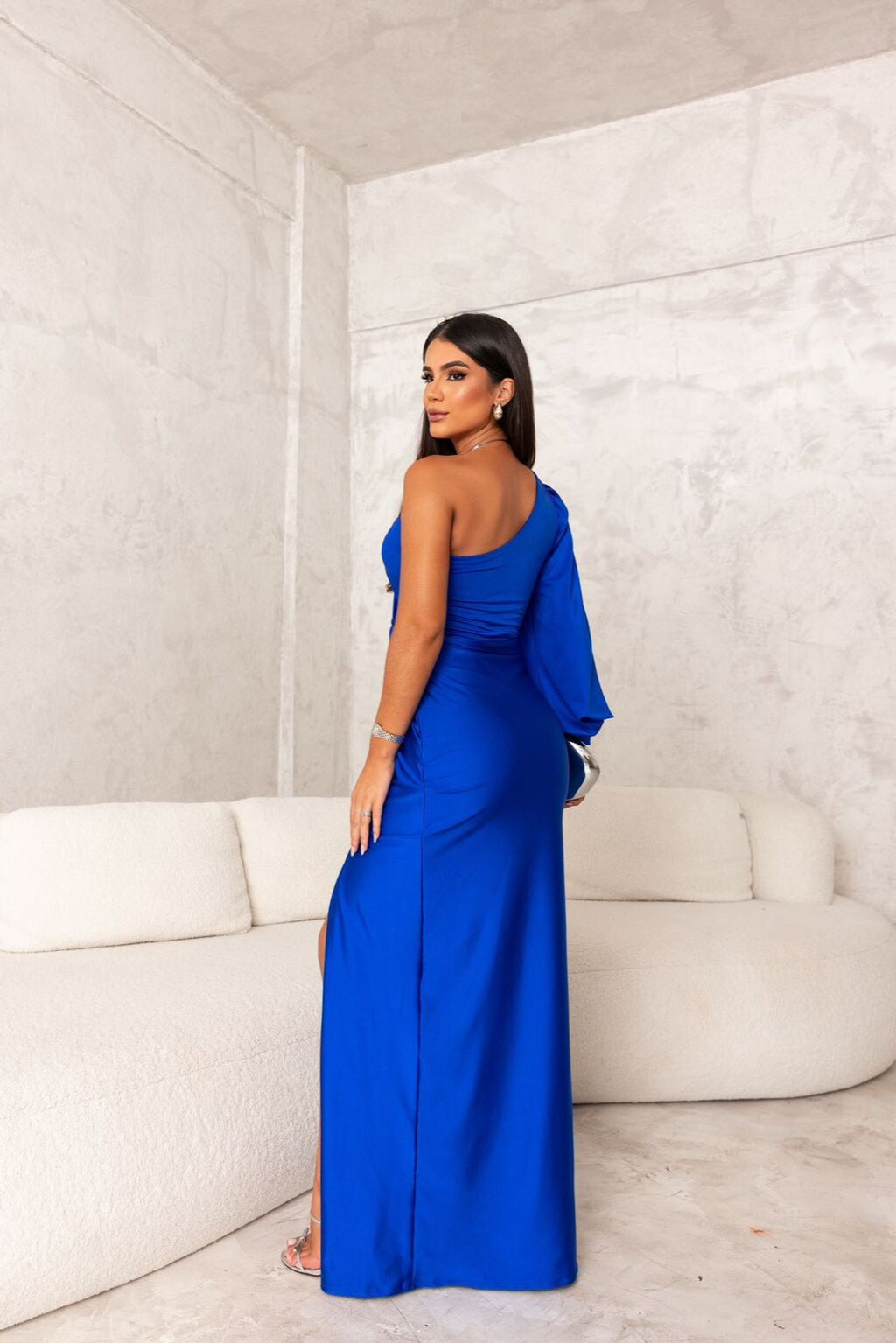 Donna in abito lungo blu elegante, monospalla, con spacco laterale, in un salotto moderno con divano bianco. Moda femminile, abbigliamento da sera.