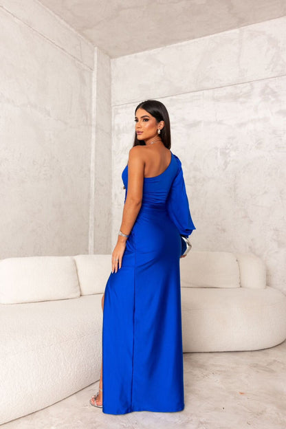Donna in abito lungo blu elegante, monospalla, con spacco laterale, in un salotto moderno con divano bianco. Moda femminile, abbigliamento da sera.