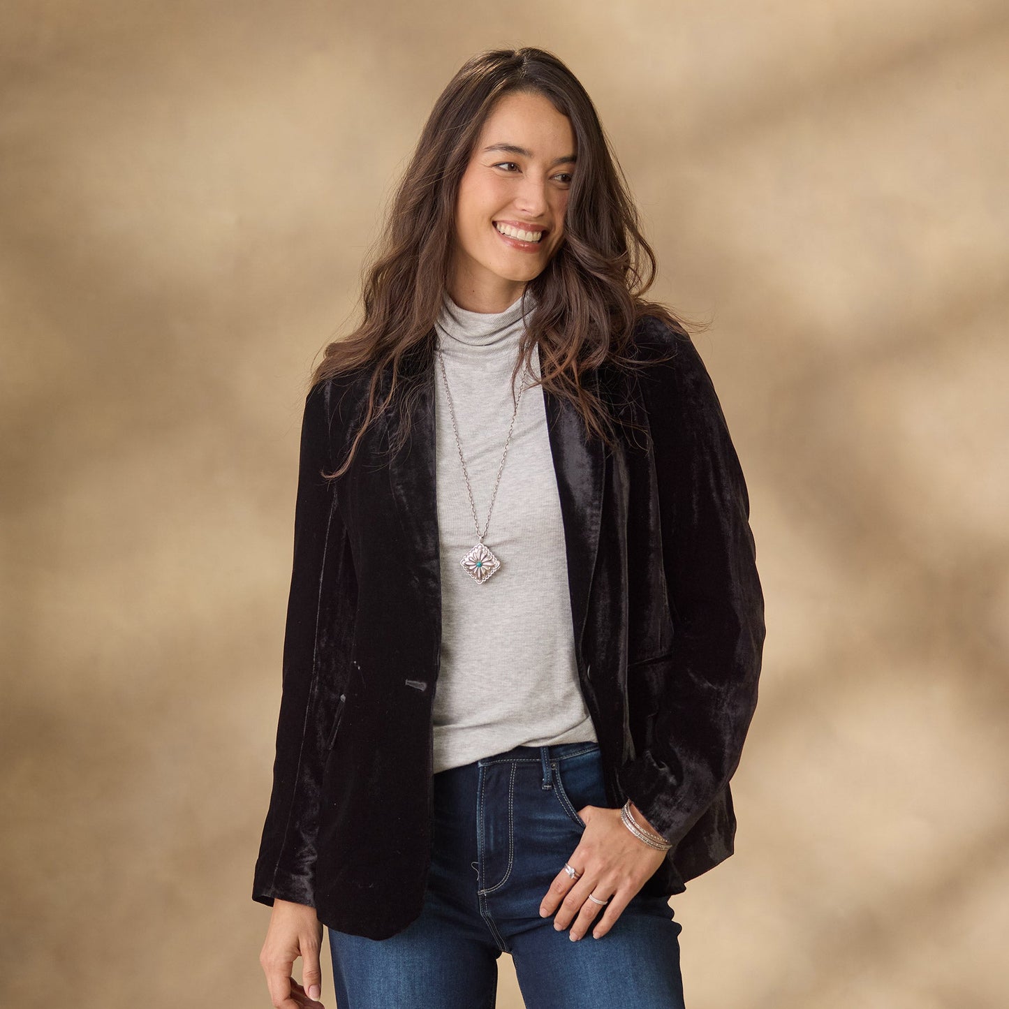 Donna sorridente con giacca di velluto nero, maglia grigia a collo alto e jeans blu. Sfondo beige sfocato. Moda casual elegante autunnale.