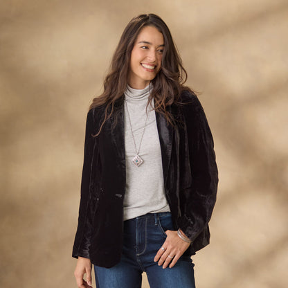 Donna sorridente con giacca di velluto nero, maglia grigia a collo alto e jeans blu. Sfondo beige sfocato. Moda casual elegante autunnale.