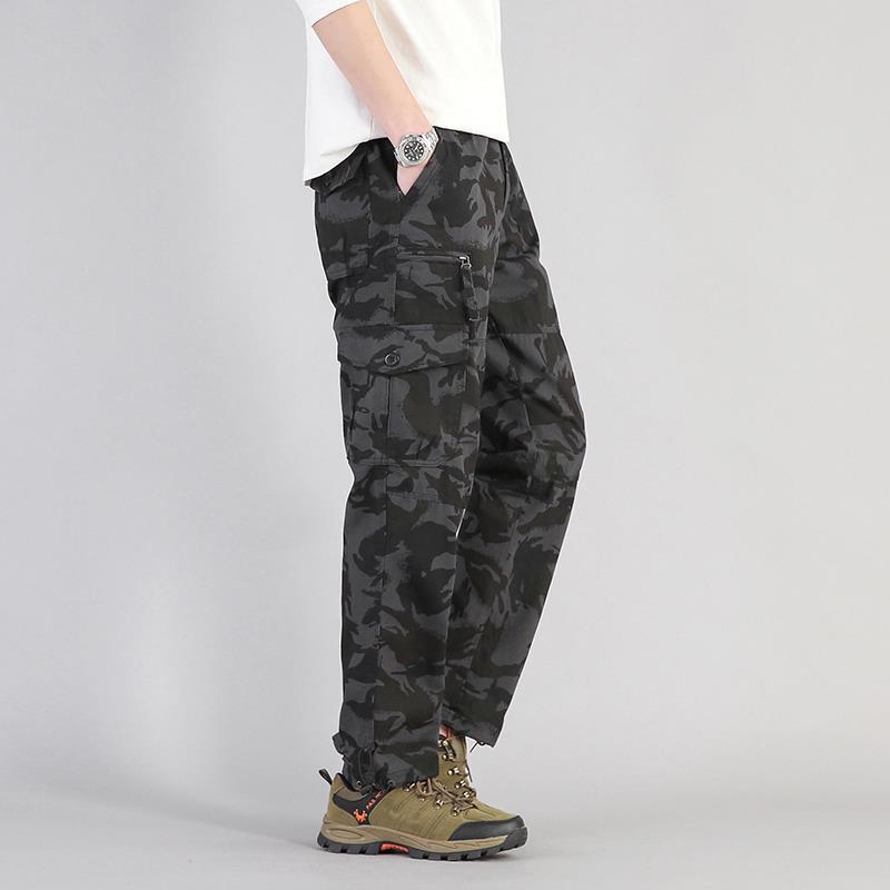 Pantaloni cargo mimetici neri da uomo, stile militare, con tasche multiple, abbinati a scarpe da trekking marroni e maglietta bianca.