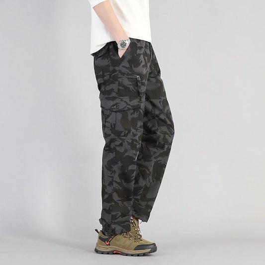 Pantaloni cargo mimetici neri da uomo, stile militare, con tasche multiple, abbinati a scarpe da trekking marroni e maglietta bianca.