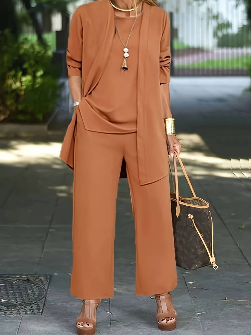 Donna in elegante completo marrone, pantaloni larghi e giacca lunga, con borsa di lusso e sandali con zeppa, passeggia in ambiente urbano.