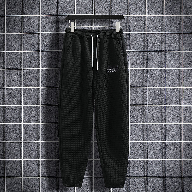 Pantaloni jogger neri a quadri appesi su gruccia, sfondo grigio a griglia. Stile casual, moda uomo, abbigliamento sportivo.