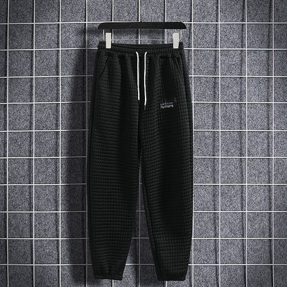 Pantaloni jogger neri a quadri appesi su gruccia, sfondo grigio a griglia. Stile casual, moda uomo, abbigliamento sportivo.