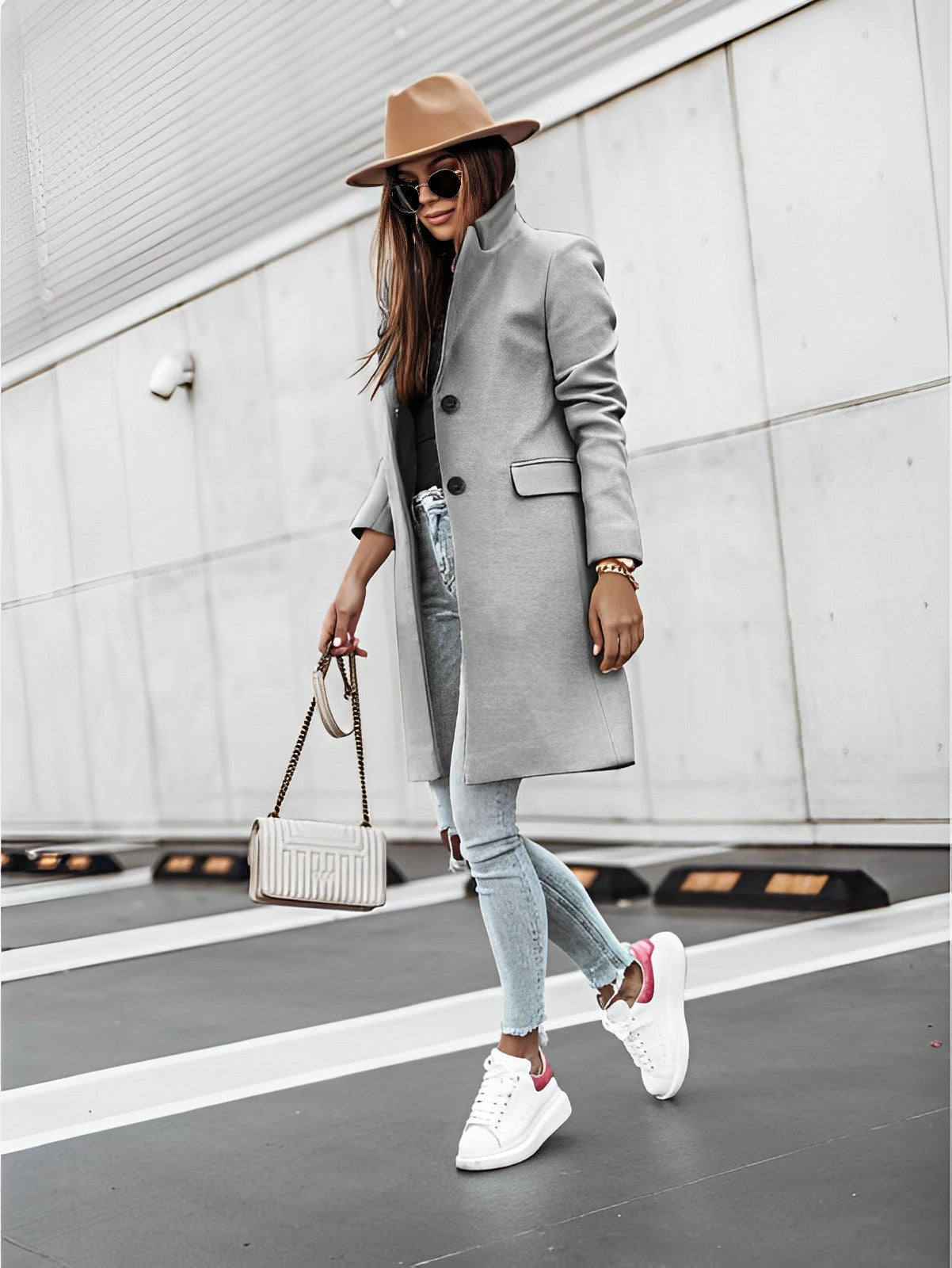 Donna alla moda con cappotto grigio, jeans strappati, sneakers bianche e cappello marrone, cammina su strada urbana. Borsa a tracolla elegante.