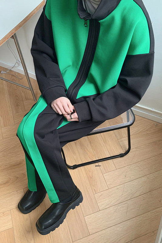 Persona seduta su sedia indossa tuta sportiva verde e nera con zip, pantaloni abbinati e stivali neri. Stile casual, moda urbana.