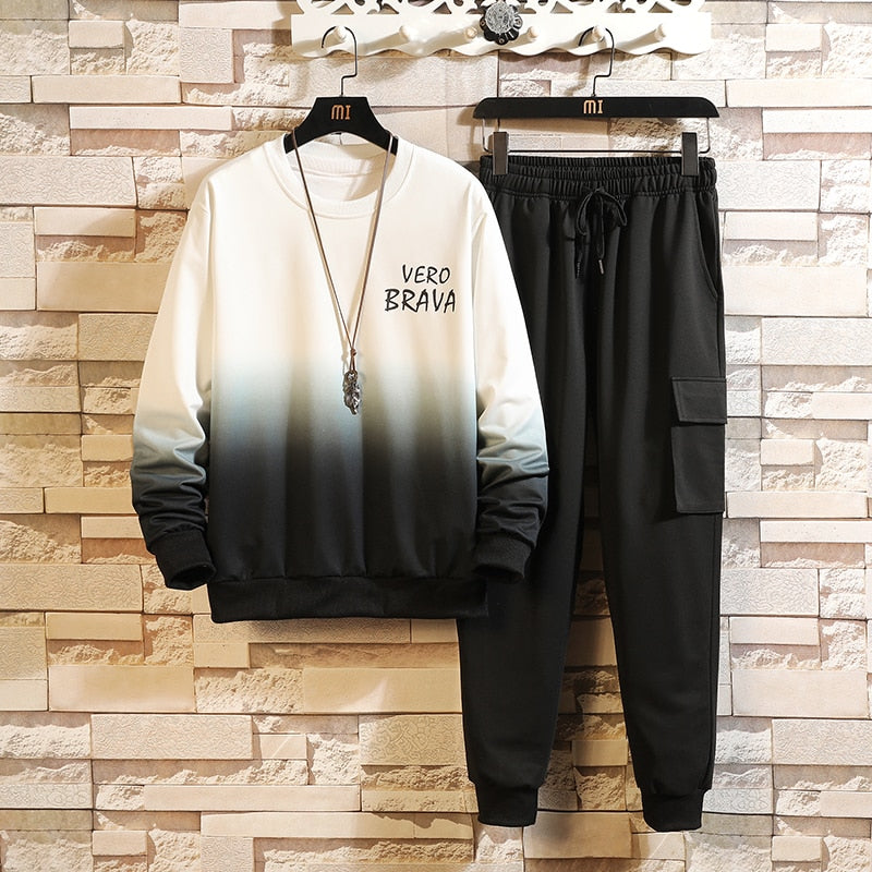 Tuta sportiva uomo bianca e nera sfumata, con scritta "Vero Brava", appesa su grucce davanti a muro in pietra. Stile casual, moda maschile.