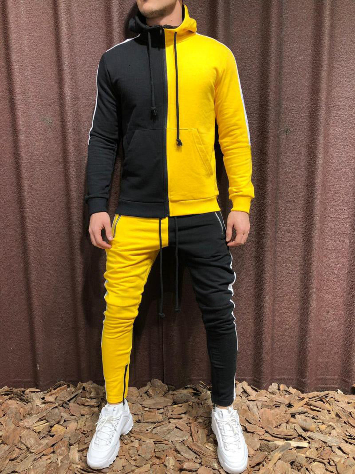 Tuta sportiva bicolore giallo e nero, uomo in piedi su sfondo marrone, scarpe bianche. Abbigliamento casual, moda streetwear.