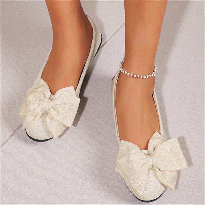 Scarpe ballerine bianche con fiocco grande, indossate su pavimento chiaro. Eleganti e alla moda, perfette per look casual e raffinati.