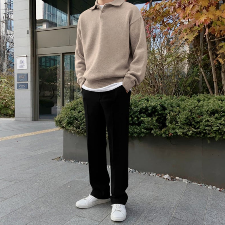 Uomo in abbigliamento casual autunnale: maglione beige, pantaloni neri, scarpe bianche. Sfondo urbano con piante e edificio moderno.