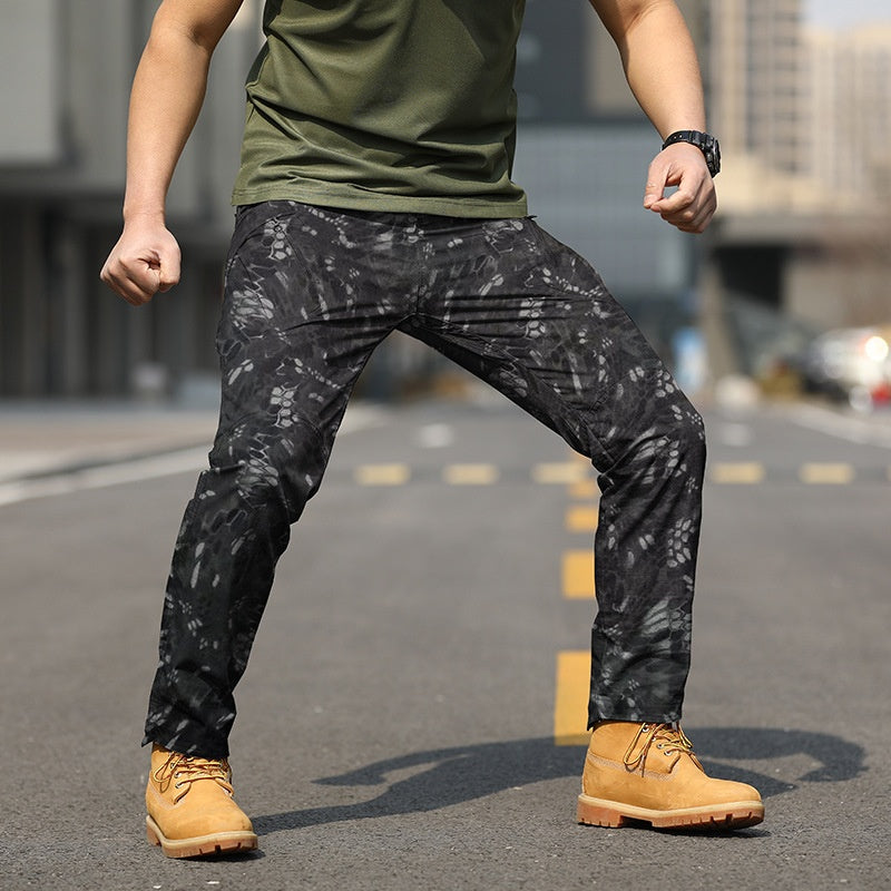 Uomo in posa su strada indossa pantaloni mimetici neri e stivali gialli, sfondo urbano sfocato. Moda maschile, abbigliamento casual.