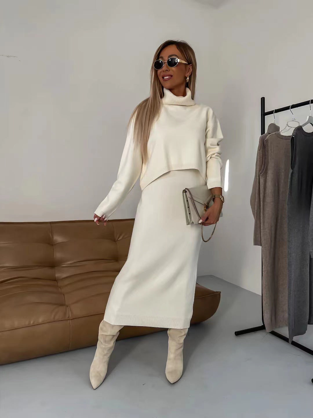 Donna con abito bianco in maglia, stivali beige e occhiali da sole, accanto a divano marrone e stand con abiti. Moda autunnale elegante.