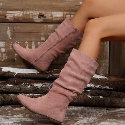 Stivali rosa in pelle scamosciata con cerniera laterale, indossati da una persona seduta su un fondo di legno rustico. Moda autunnale elegante.