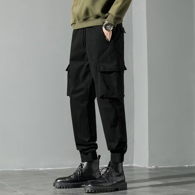 Pantaloni cargo neri da uomo, stile casual, con tasche laterali, abbinati a stivali neri robusti e maglione verde oliva. Moda maschile autunnale.