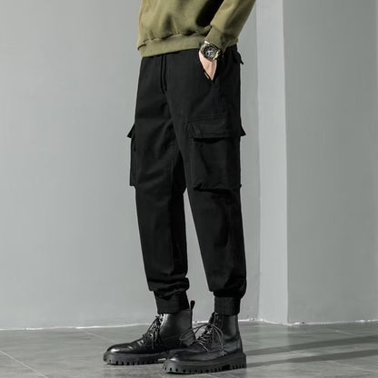 Pantaloni cargo neri da uomo, stile casual, con tasche laterali, abbinati a stivali neri robusti e maglione verde oliva. Moda maschile autunnale.