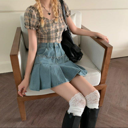 Giovane donna seduta su sedia in legno, indossa top a quadri, gonna di jeans, calze bianche in pizzo e stivali neri. Stile moda casual chic.