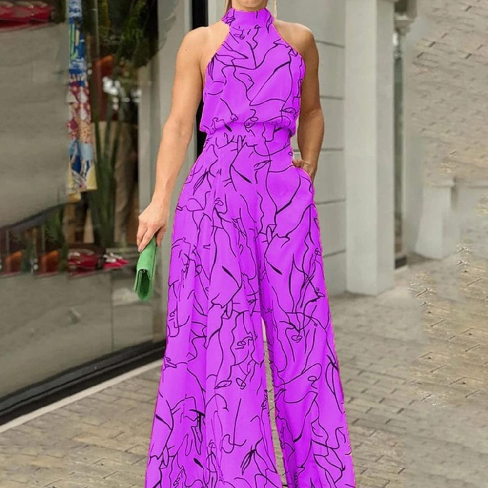Tuta elegante viola con motivo astratto nero, indossata da una donna in piedi su un marciapiede. Accessorio: pochette verde. Moda estiva chic.