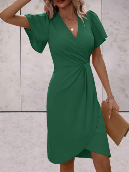 Donna in abito verde elegante con maniche a volant, indossa collana e bracciale, tiene una pochette beige. Moda femminile, abbigliamento chic.