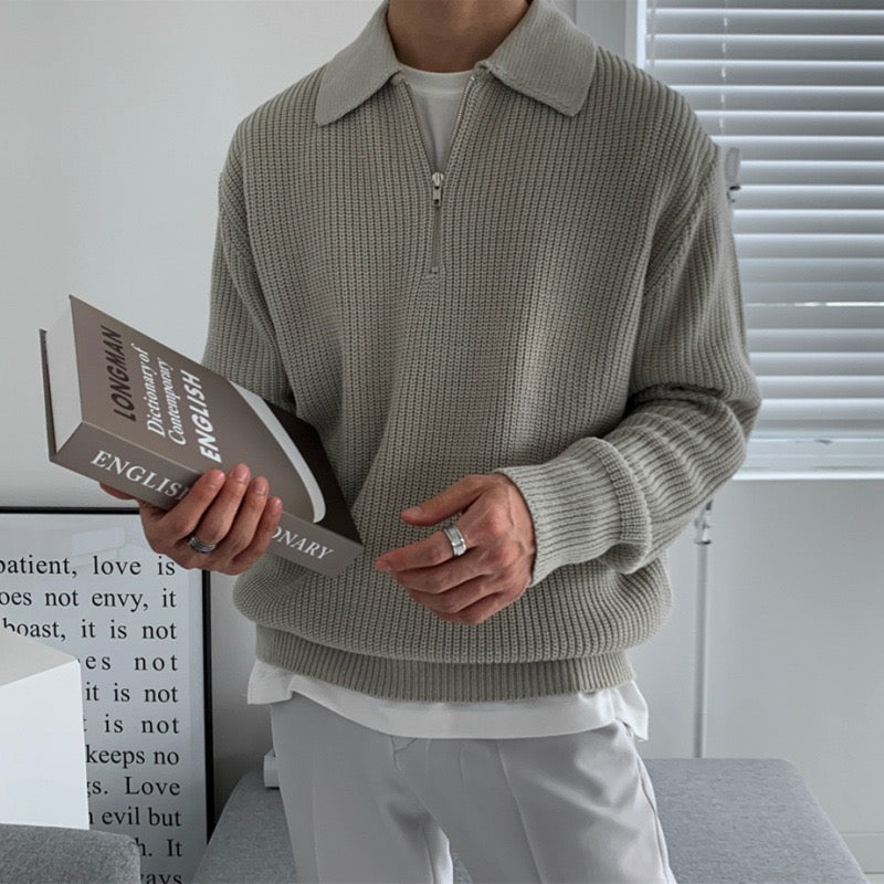 Uomo con maglione grigio a collo alto, tiene un dizionario inglese. Interno moderno, finestra con persiane. Moda casual, lettura, arredamento minimalista.