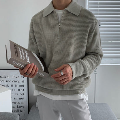 Uomo con maglione grigio a collo alto, tiene un dizionario inglese. Interno moderno, finestra con persiane. Moda casual, lettura, arredamento minimalista.