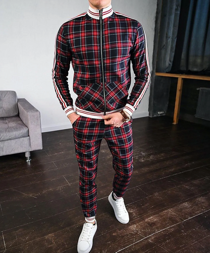 Tuta sportiva a quadri rossi e neri, uomo in piedi su pavimento in legno, scarpe bianche, divano grigio sullo sfondo. Moda casual maschile.